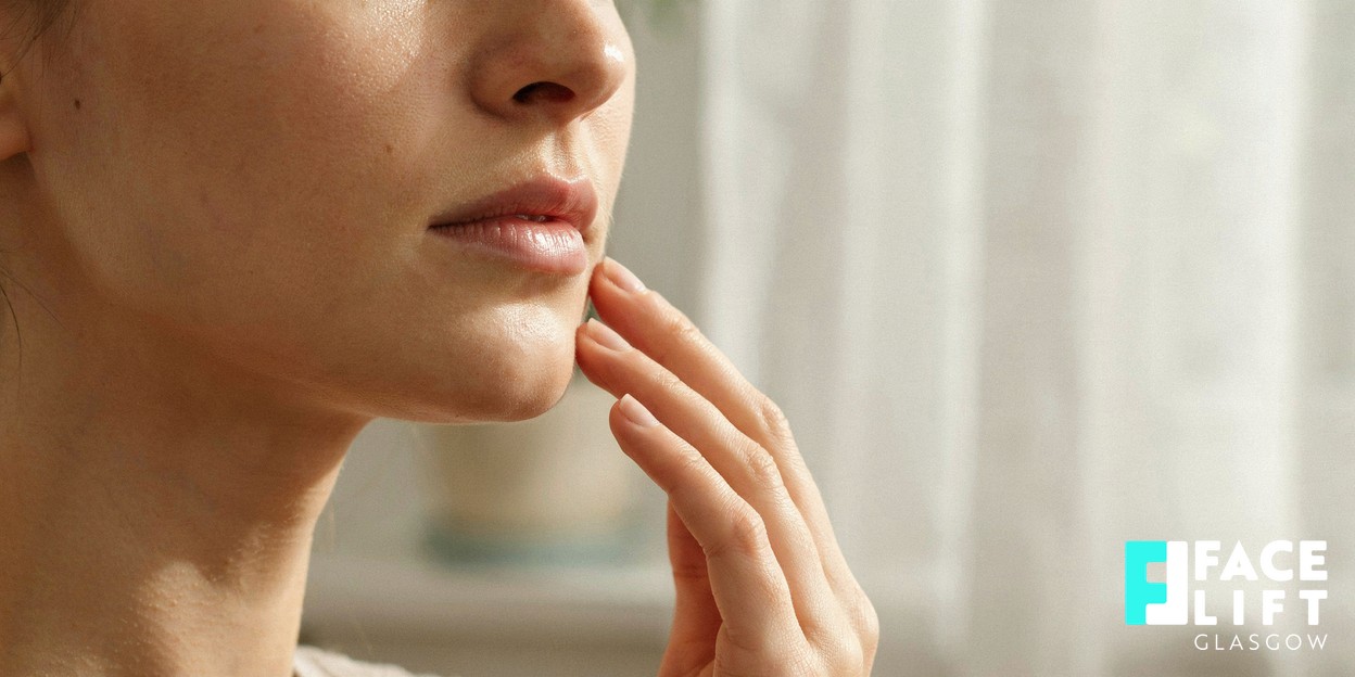 Soft-focus editorial close-up crop of a woman’s lower face and jawline, her fingertips resting lightly along the jaw as if checking skin firmness, natural window light